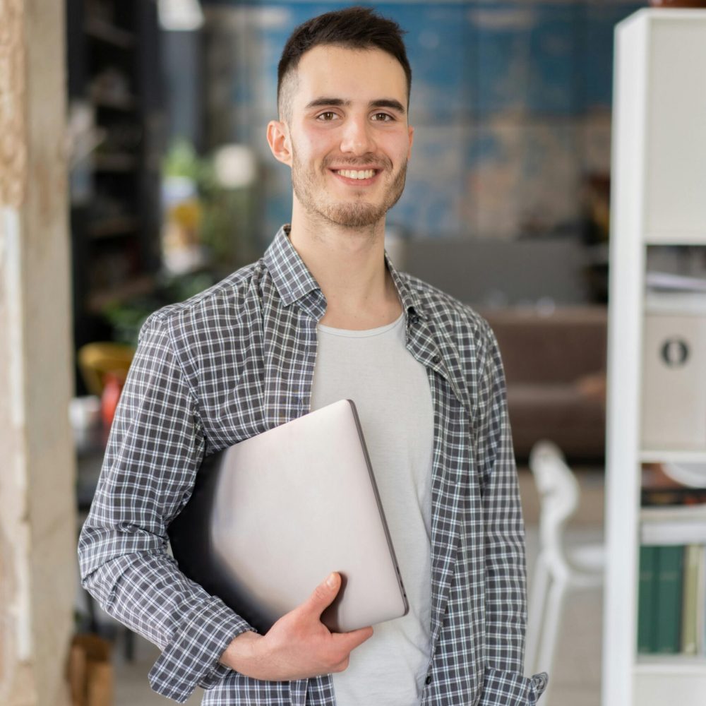 Cheerful young adult man holding a laptop, standing in a contemporary office space.